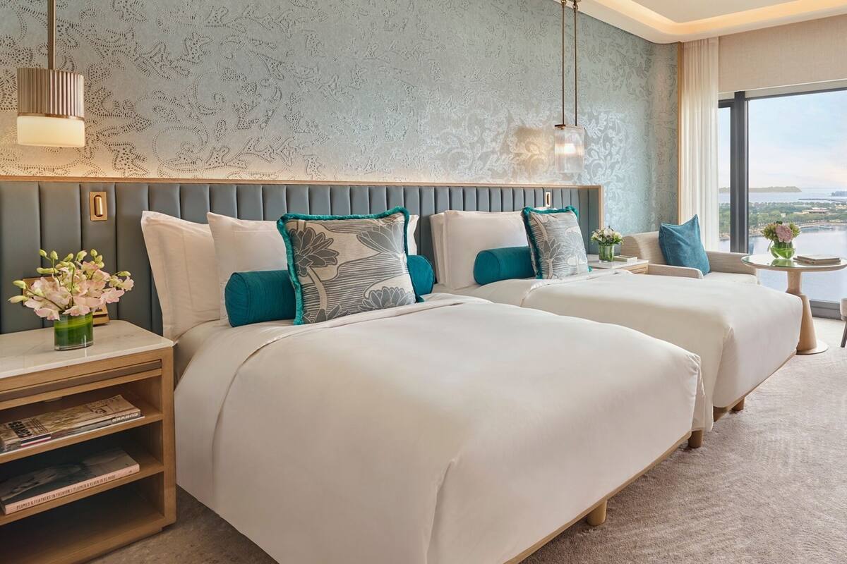 Hotelkamer met twee bedden, een nachtkastje met boeken, een klein tafeltje met een vaas en een groot raam met uitzicht.