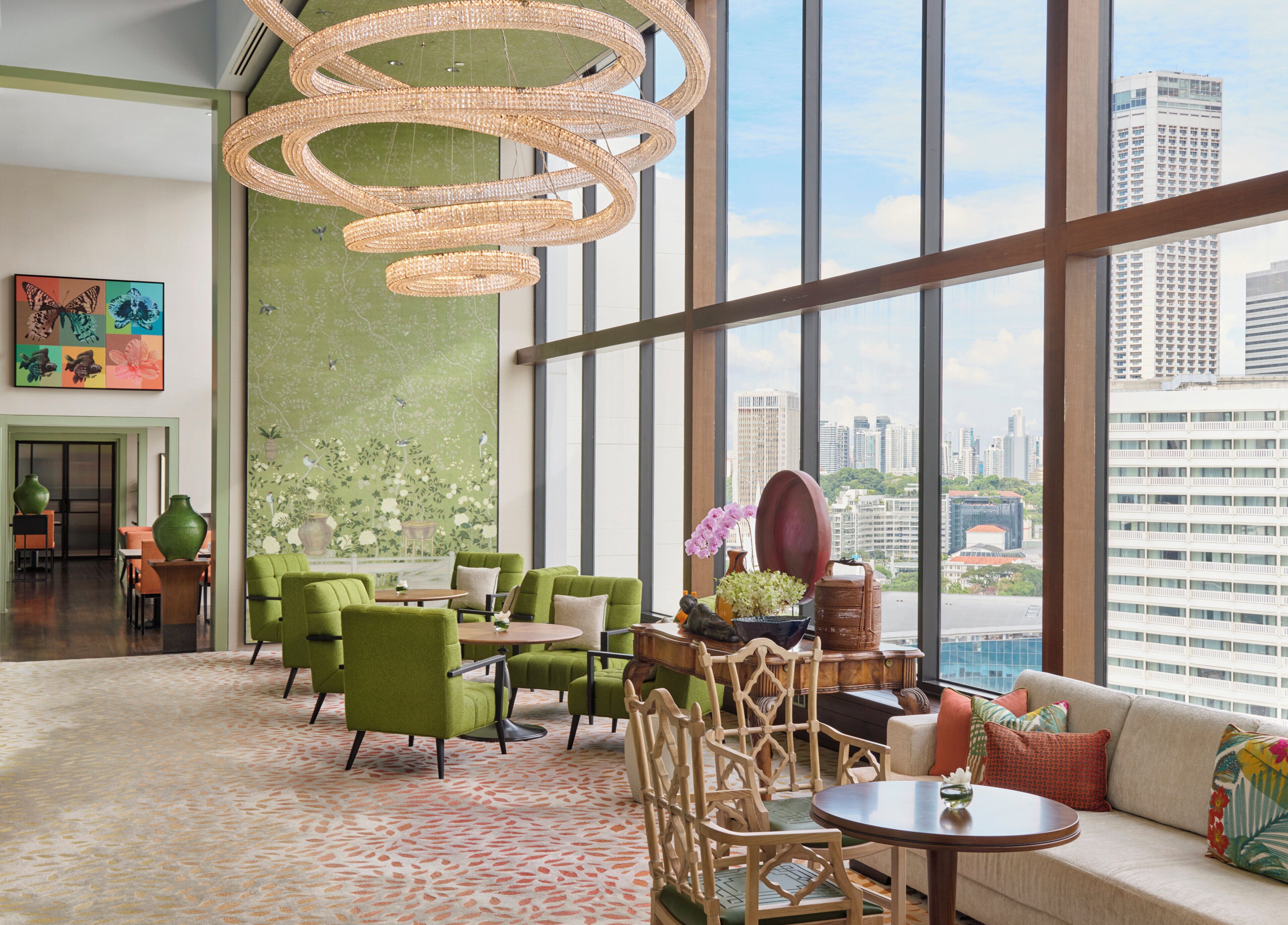 Un hall d’entrée spacieux avec un plafond haut, de grandes fenêtres offrant une vue sur la ville, des sièges recouverts d’un tissu vert et un lustre décoratif.