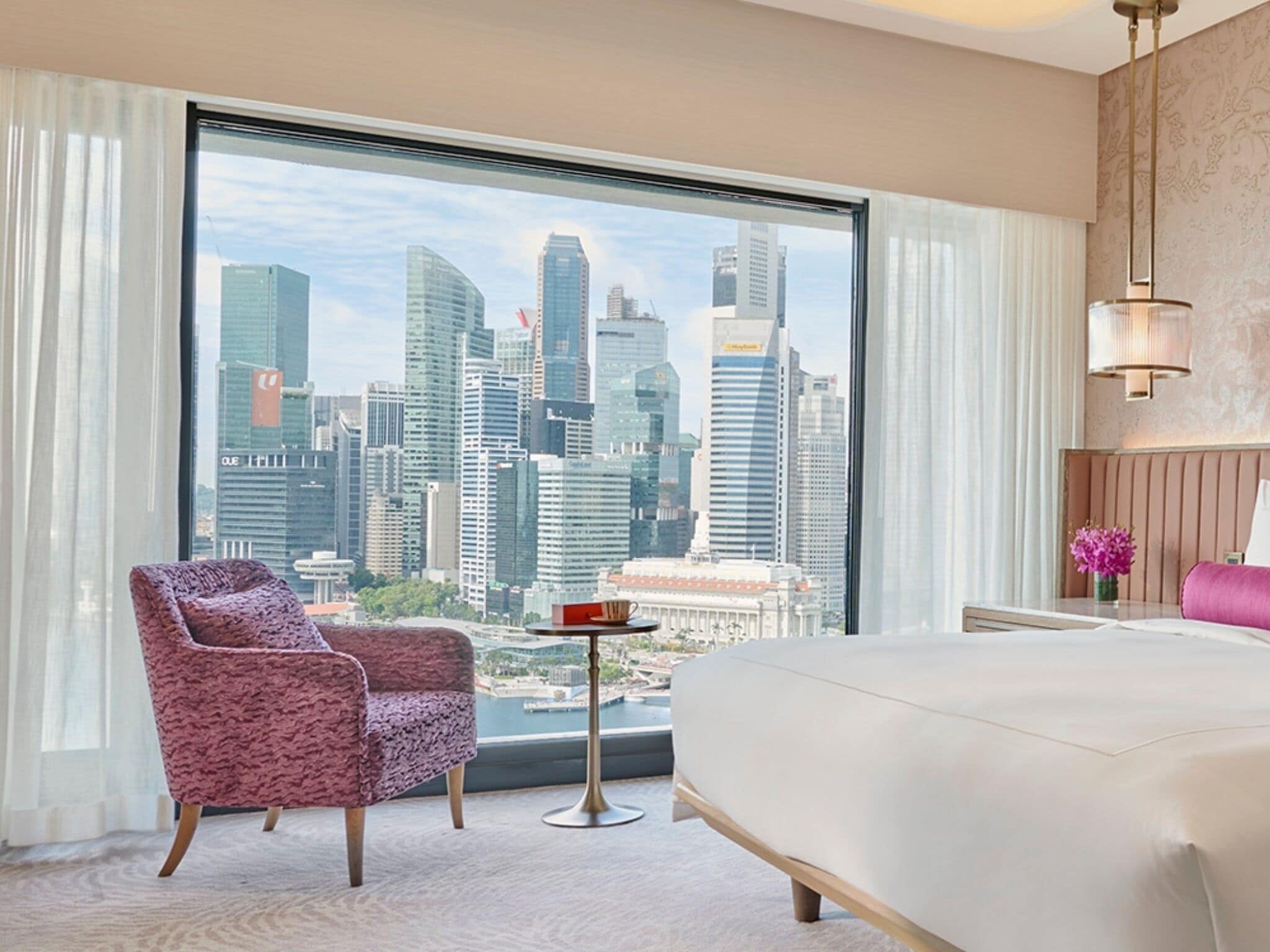 A hotel room with a large window offering a city skyline view, a bed, a chair, and a small table.