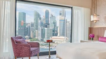 A hotel room with a large window offering a city skyline view, a bed, a chair, and a small table.