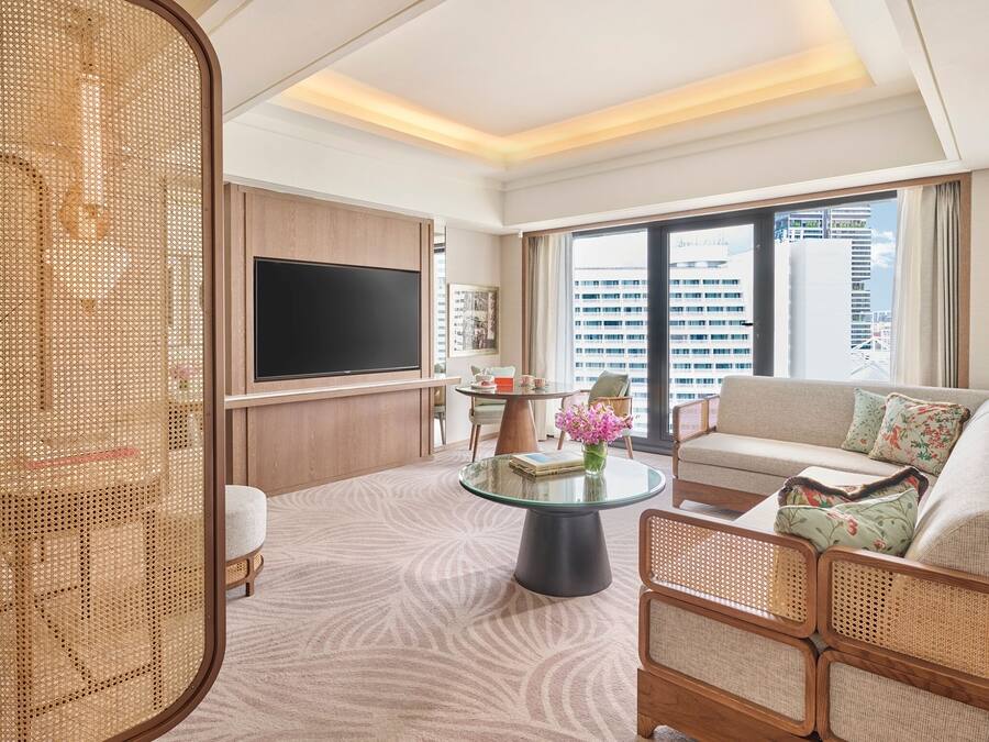 A modern hotel room with a large TV, comfortable seating, a glass coffee table, and a view of the cityscape.