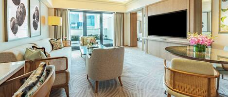 Habitación de hotel con televisor grande, zona de comedor, sofá y balcón con vistas.