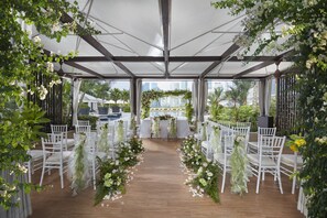 An outdoor wedding venue with white chairs and tables, floral decorations, and a view of the city skyline.