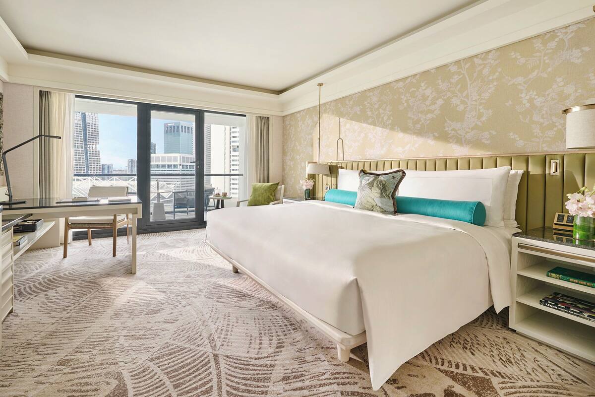 Habitación de hotel con una cama grande, un escritorio, una silla, una estantería y vistas a la ciudad.