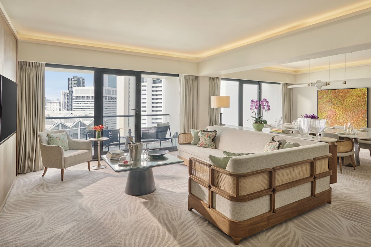 Een ruime hotelkamer met een groot raam en uitzicht op de stad, een comfortabele zithoek, een eethoek en een modern kunstwerk aan de muur.
