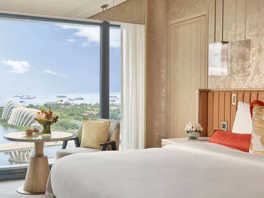 Habitación de hotel con un ventanal que ofrece vistas a la costa, una cama, una mesita de noche y una pequeña mesa con flores.