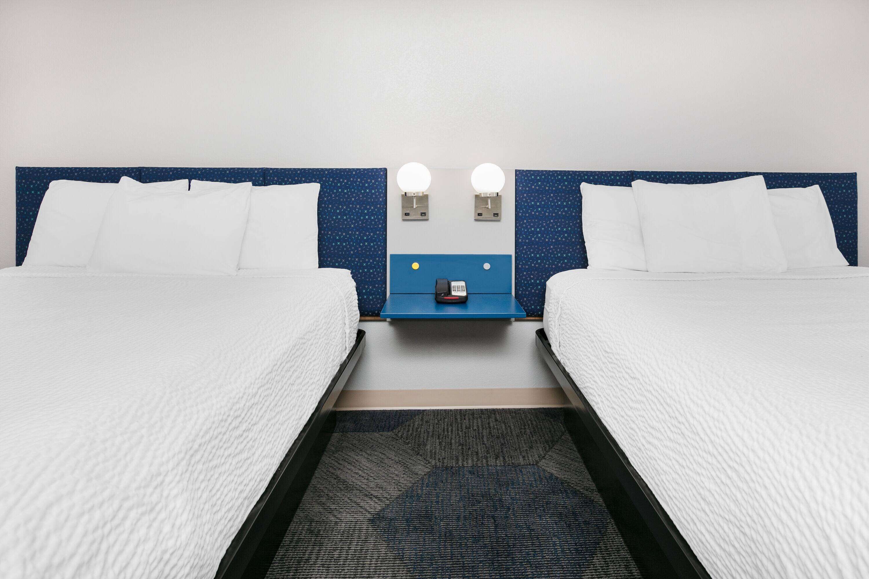A hotel room with two beds, a blue headboard, two bedside lamps, and a small table with a phone.