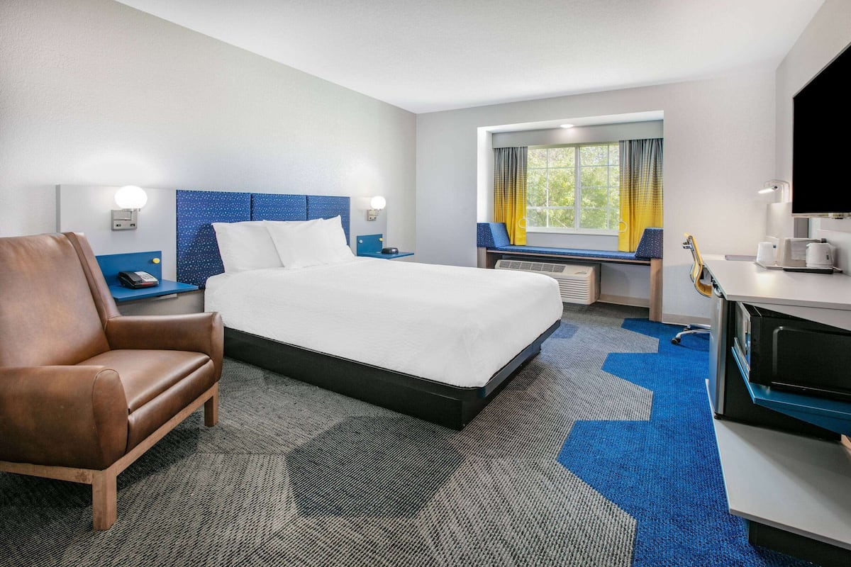 Ein Hotelzimmer mit einem Bett, einem Ledersessel, einem Schreibtisch mit Computer, einem Fenster mit Vorhängen und einem Fernseher.