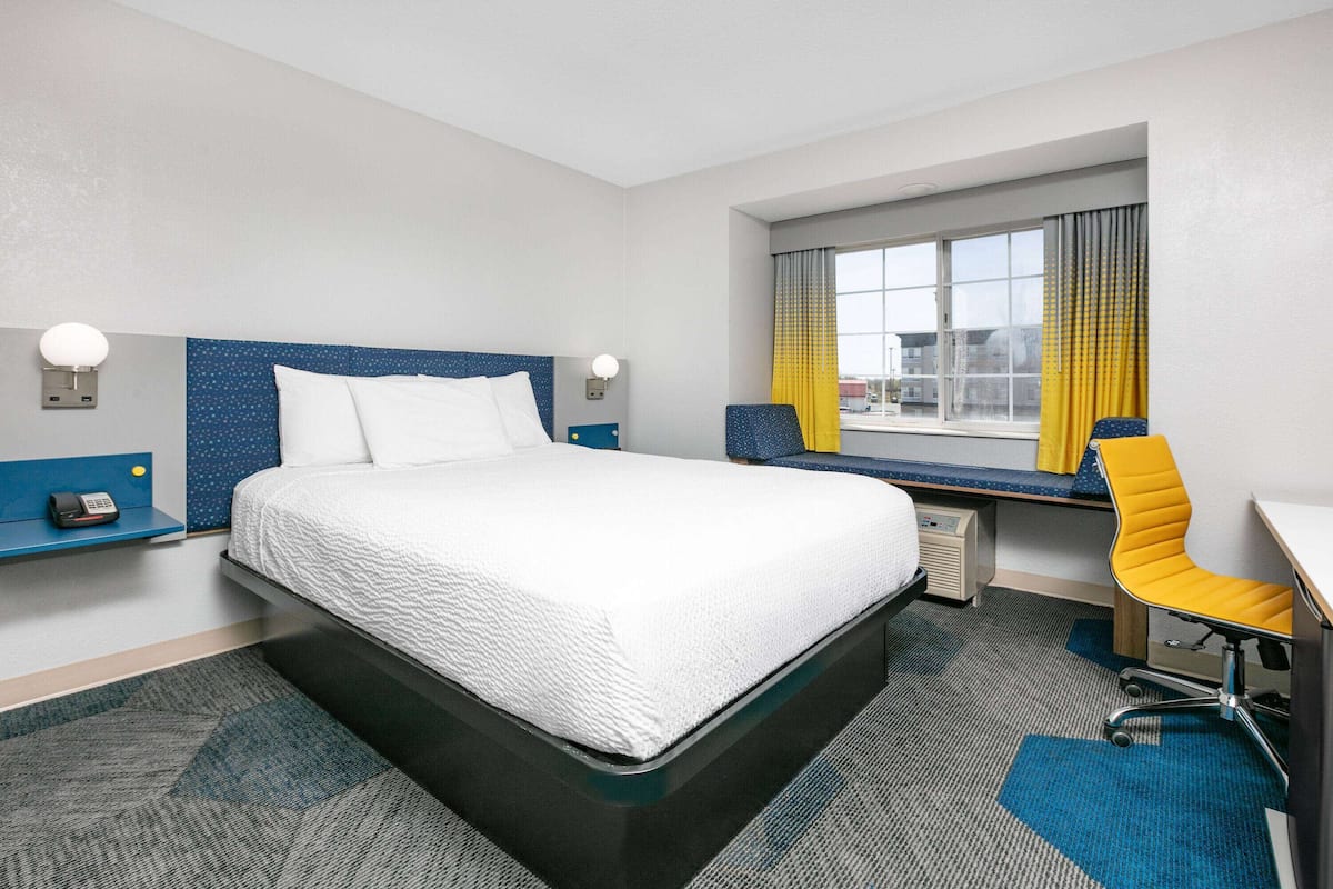 Ein Hotelzimmer mit einem großen Bett, einem Schreibtisch, einem Stuhl, einem Fenster mit Vorhängen und einem Telefon auf dem Nachttisch.