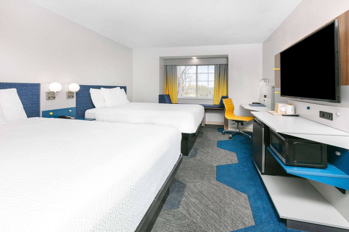 Ein Hotelzimmer mit zwei Betten, einem Schreibtisch, einem Fernseher und einem Fenster mit Vorhängen.