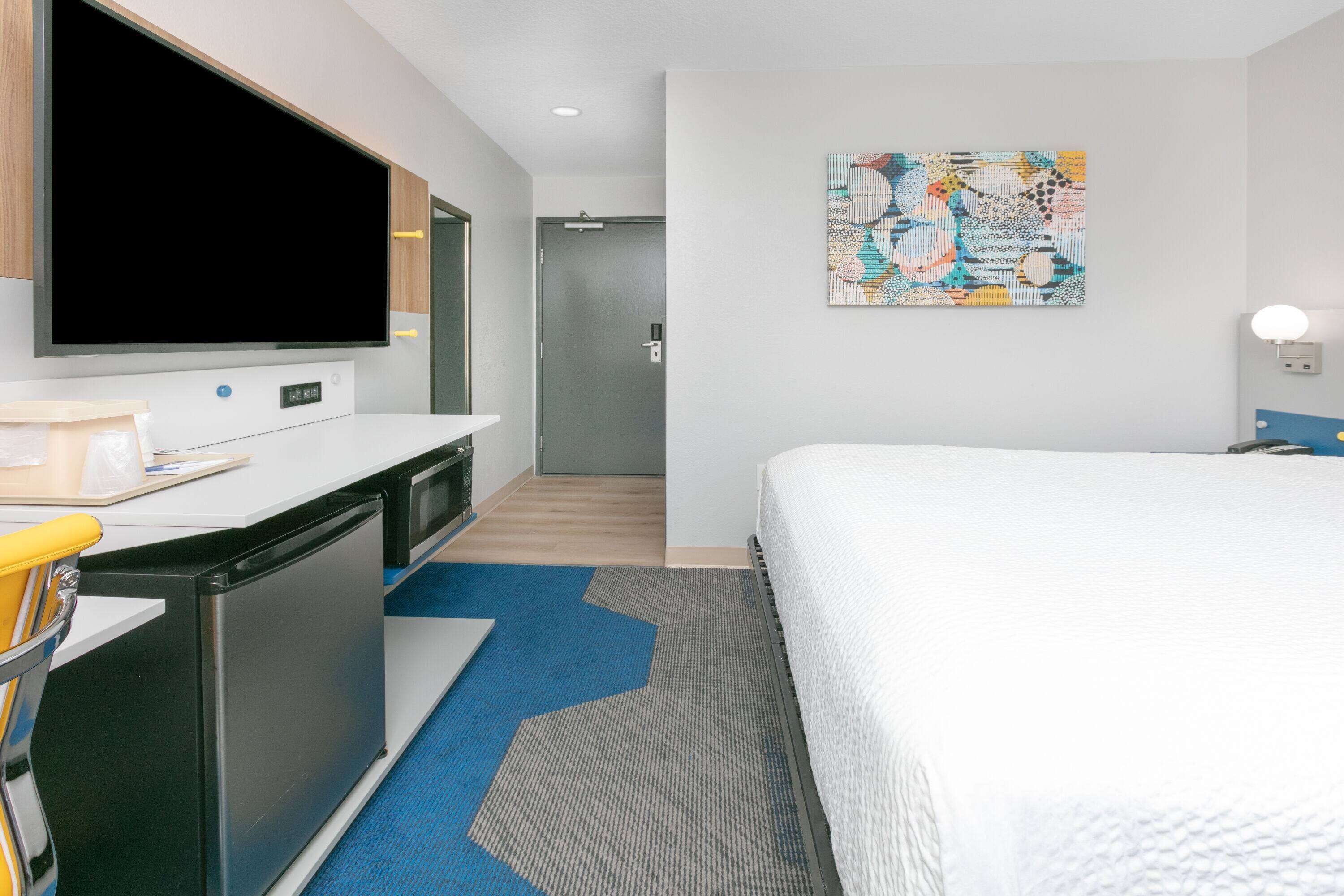 A hotel room with a bed, a desk with a flat-screen TV, a desk lamp, and a colorful abstract painting on the wall.