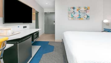 A hotel room with a bed, a desk with a flat-screen TV, a desk lamp, and a colorful abstract painting on the wall.
