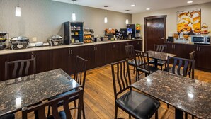 A restaurant with dark wooden tables and chairs, a counter with various food items and a coffee machine, and a wall with a picture of food. - Best Western Crown Inn & Suites (Batavia)