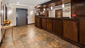 A reception area with a wooden reception desk, a small table with a vase of flowers, and a tiled floor. - Best Western Crown Inn & Suites (Batavia)