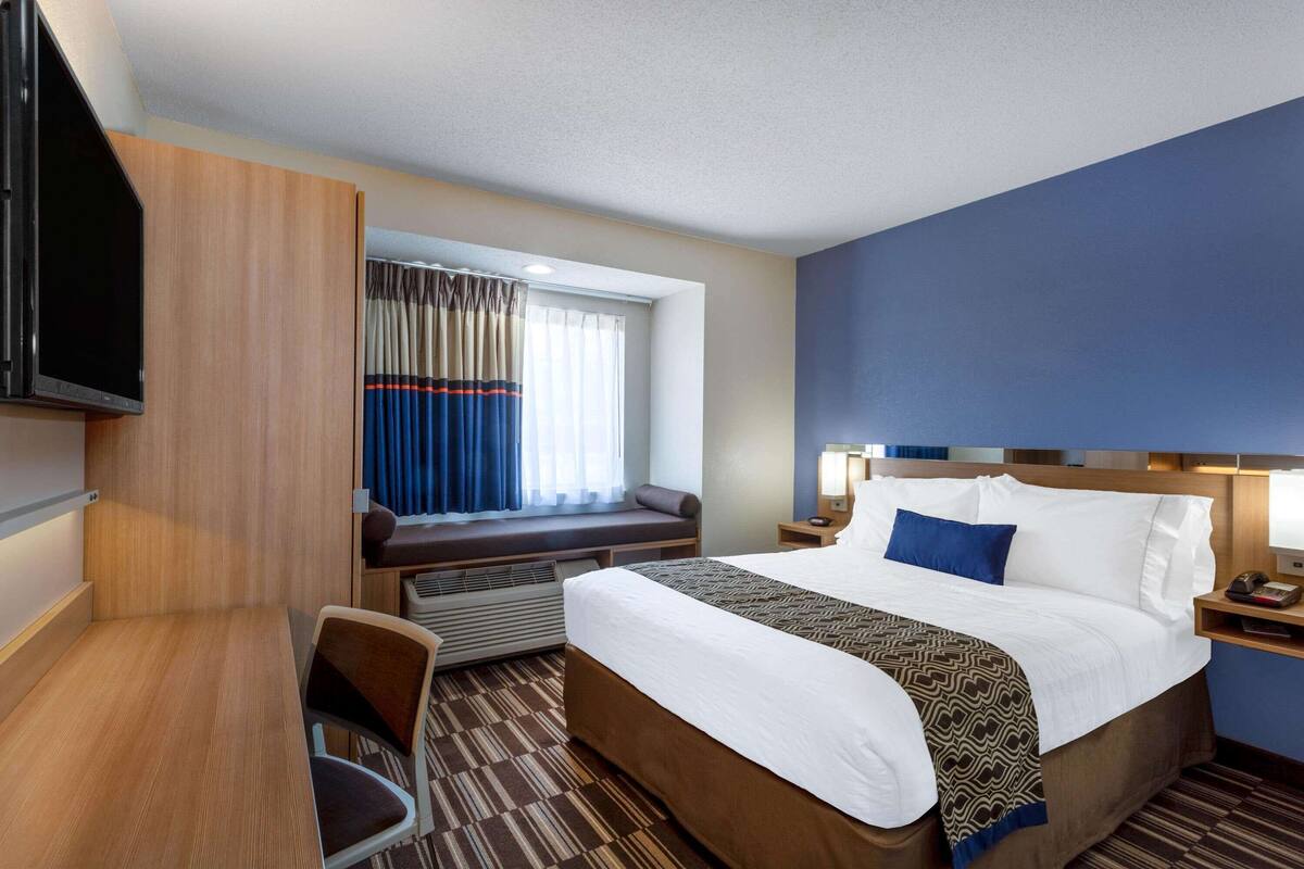 A hotel room with a bed, a desk, a chair, a TV, a window with curtains, and a nightstand with a lamp.