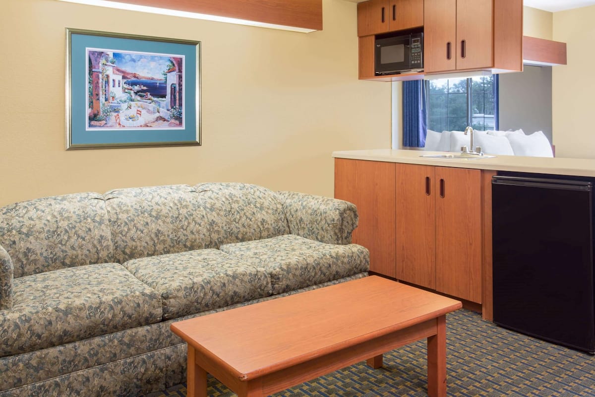 A room with a sofa, a wooden table, a kitchen area with a microwave, and a framed picture on the wall.