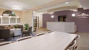 A hotel reception area with a long reception desk, seating area with chairs and a round table, and a large window with blinds. - Microtel Inn & Suites by Wyndham Leesburg/Mt Dora (Leesburg)
