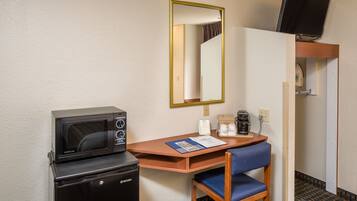 A compact hotel room with a kitchenette, a single bed, a small desk with a chair, a mirror, and a television mounted on the wall.