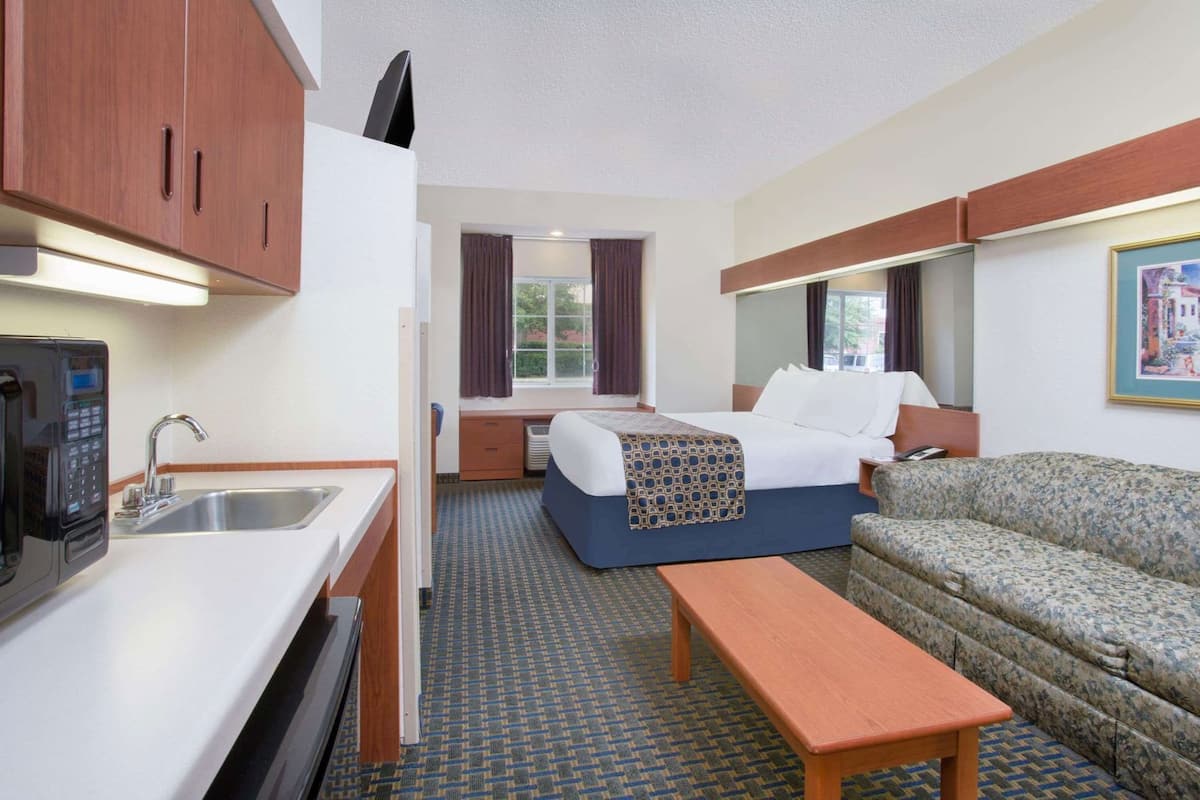 A compact hotel room with a kitchenette, a bed, a sofa, and a window.