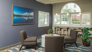 A room with a checkered floor, two armchairs, a round table, a potted plant, and a painting of a lake. - Microtel Inn & Suites by Wyndham Leesburg/Mt Dora (Leesburg)