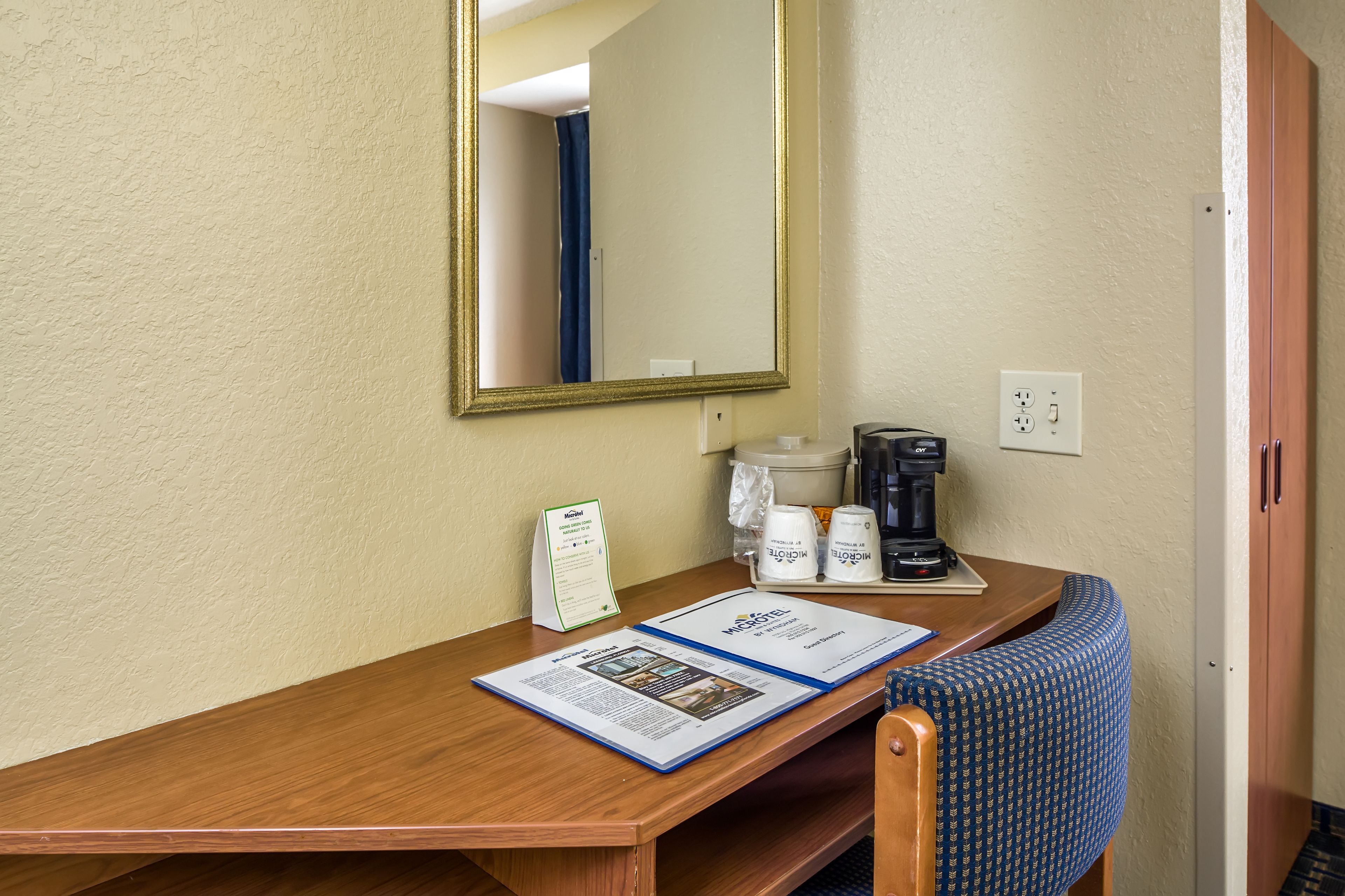 A hotel room with a wooden desk, a mirror, a coffee maker, a telephone, and a brochure on the desk.
