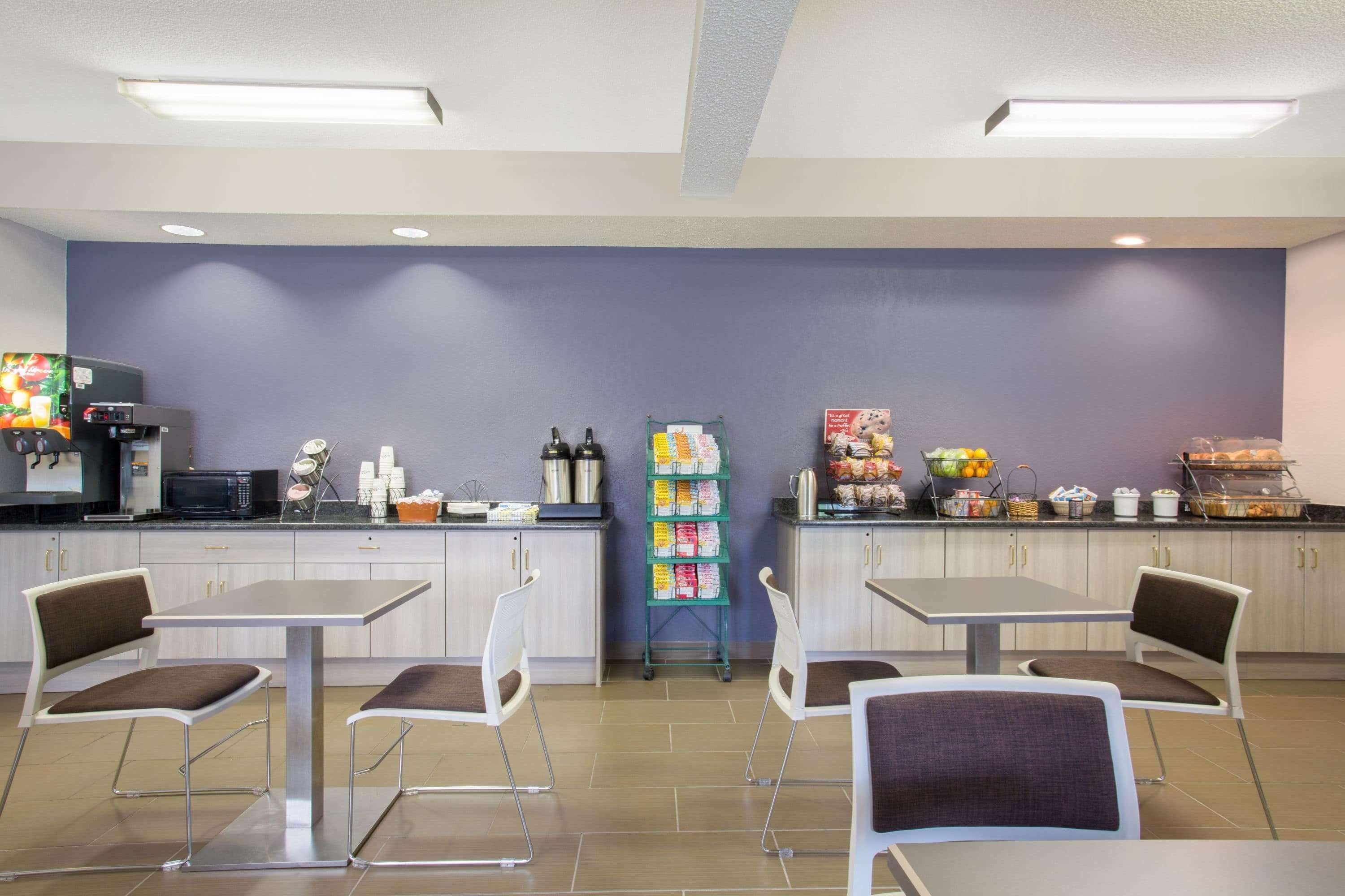 A modern cafeteria with two tables and four chairs, a microwave, beverage dispensers, and a snack bar.