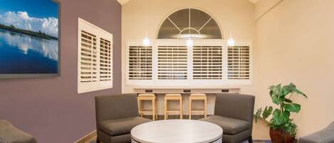 A room with a round table, two sofas, a large window with shutters, and a potted plant.