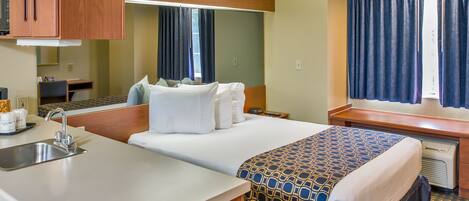 A hotel room with a bed, a small desk, a mirror, a window with curtains, a sink, and a microwave.