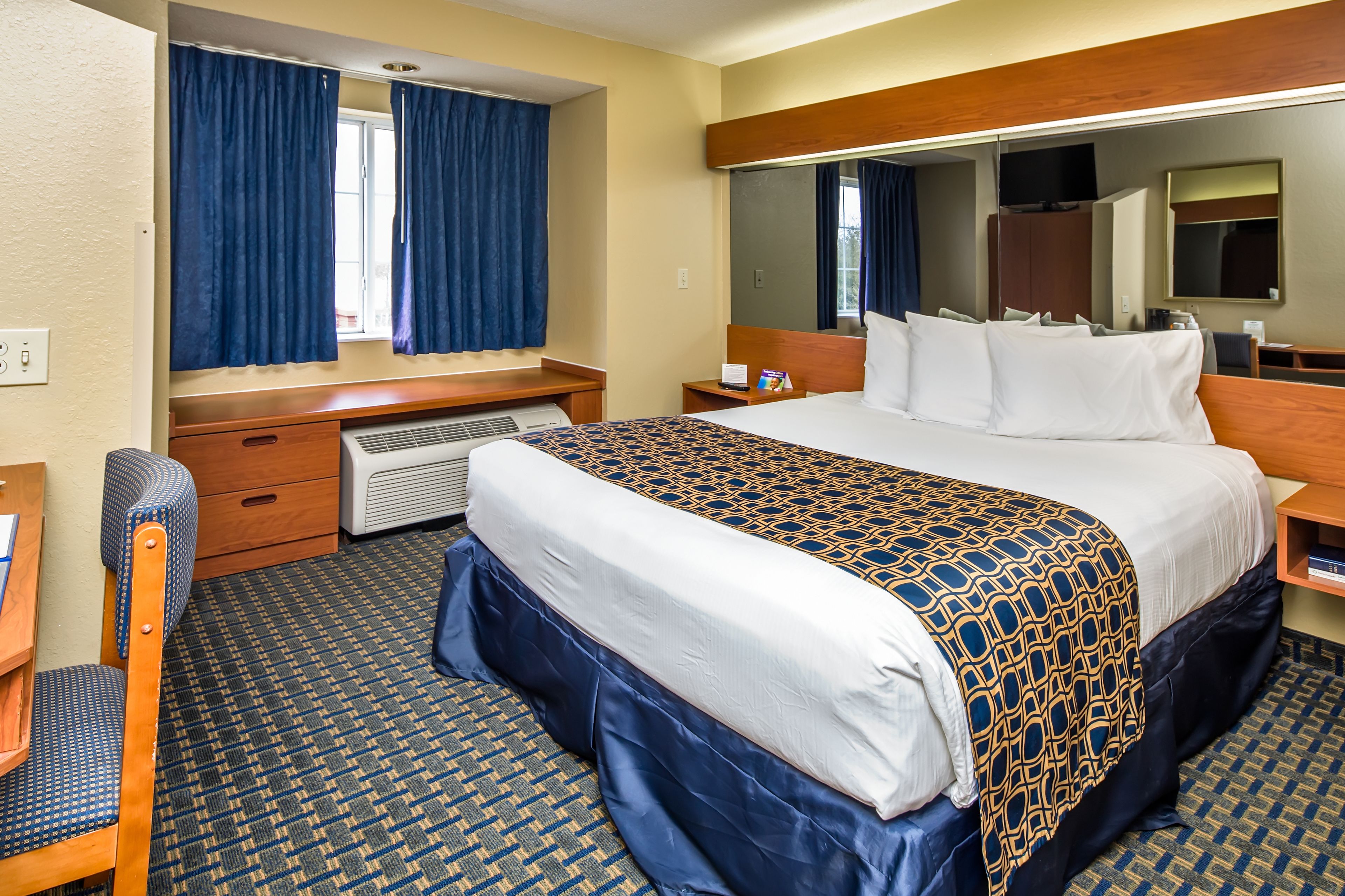 A hotel room with a large bed, a desk, a chair, a dresser, a mirror, and a window with curtains.