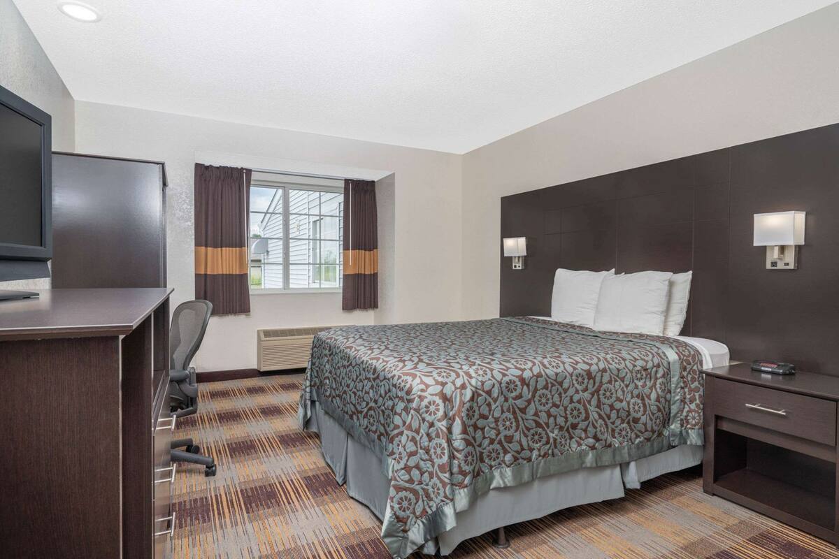 A hotel room with a bed, a desk, a chair, a TV, a window with curtains, and a wall-mounted lamp.