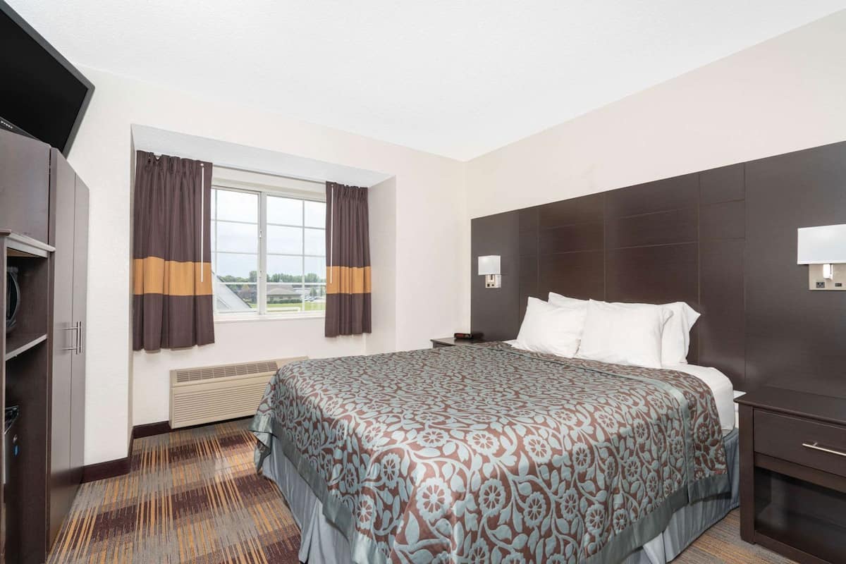A hotel room with a large bed, a TV, a window with curtains, a nightstand, and a wall-mounted lamp.