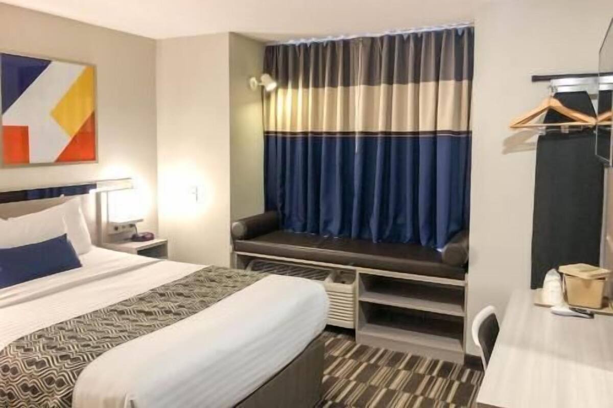 Ein Hotelzimmer mit Bett, Nachttisch, Lampe, Fenster mit Vorhängen und Schreibtisch mit Stuhl.