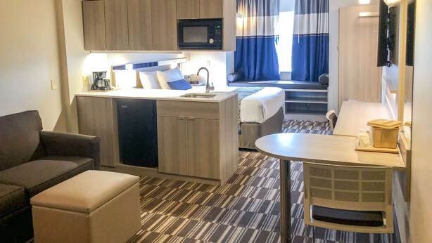 Habitación de hotel compacta con kitchenette, sofá, pequeña mesa de comedor y vista al dormitorio contiguo.