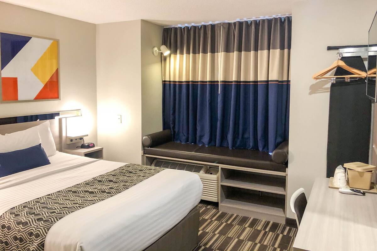 Ein Hotelzimmer mit Bett, Nachttisch, Lampe, Schreibtisch, Stuhl und einem Fenster mit Vorhängen.