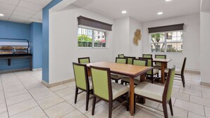 Un comedor con una mesa rectangular rodeada de sillas, un mostrador tipo buffet y ventanas con persianas.
