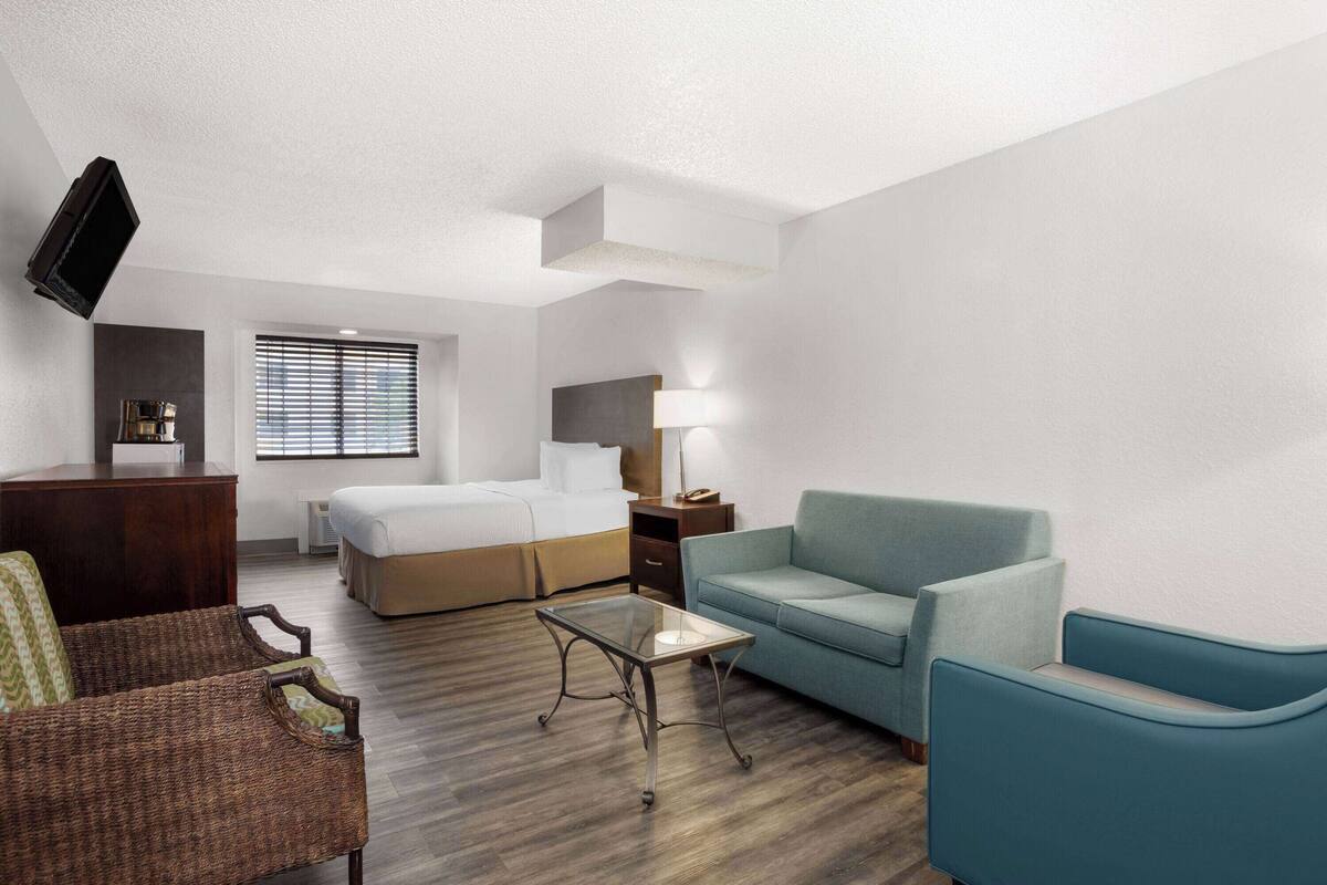 A hotel room with a bed, a sofa, a small table, a TV, and a window with blinds.