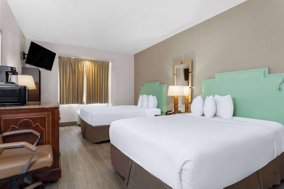 A hotel room with two beds, a desk with a chair, a TV mounted on the wall, and a window with curtains.