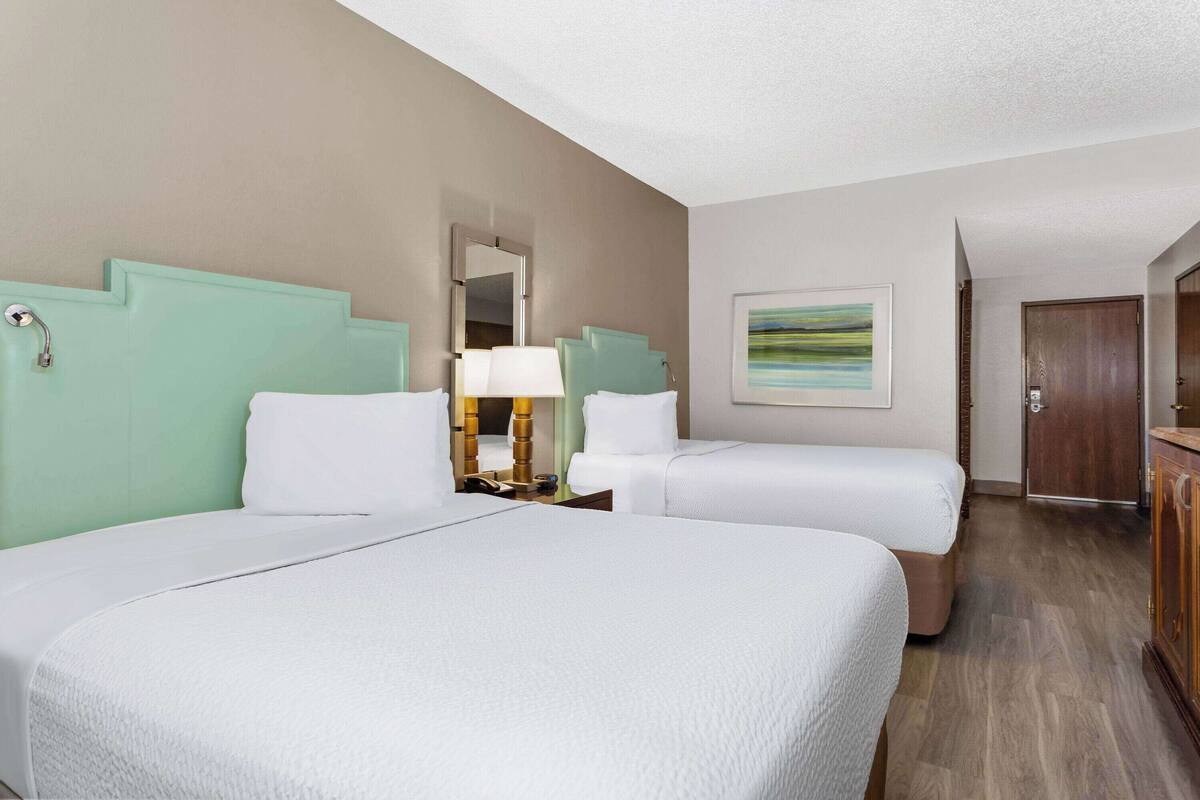 A hotel room with two beds, a nightstand with a lamp, a painting on the wall, and a wooden door.