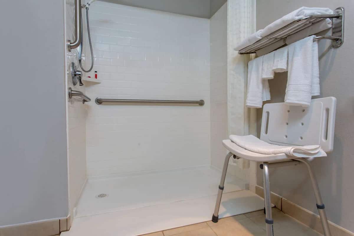 A bathroom with a white tiled shower, a grab bar, a white toilet with a safety seat, and a towel rack with towels.
