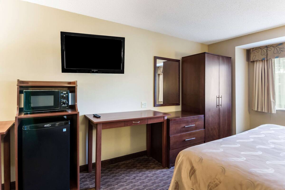 A hotel room with a bed, a desk with a microwave, a refrigerator, a wardrobe, and a flat-screen TV mounted on the wall.