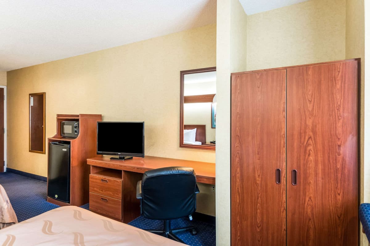 A hotel room with a bed, a desk with a computer and a TV, a wardrobe, and a small refrigerator.