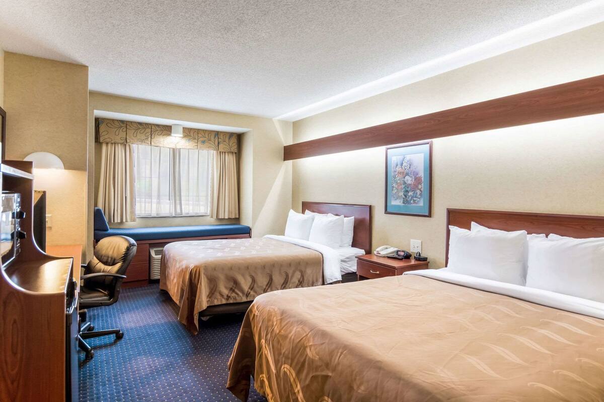 A hotel room with two beds, a desk, a chair, a window with curtains, a painting on the wall, and a telephone on the nightstand.