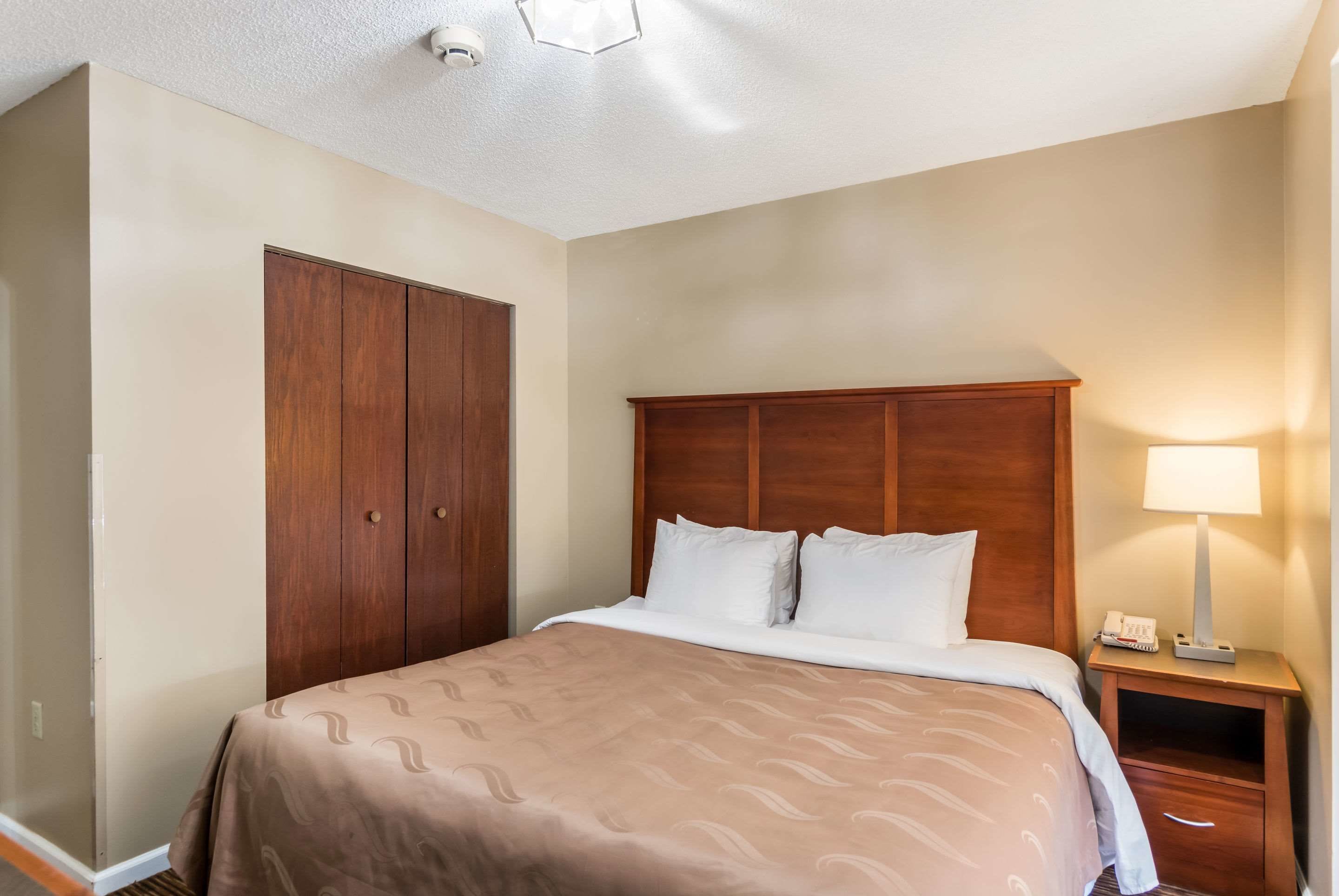 A hotel room with a large bed, a wooden wardrobe, a nightstand with a lamp, and a ceiling light.