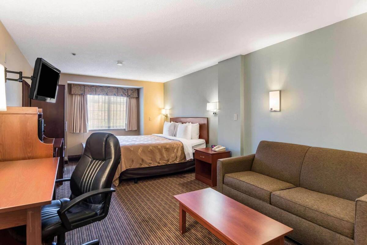 A hotel room with a bed, a desk with a chair, a sofa, a TV mounted on the wall, and a window with curtains.