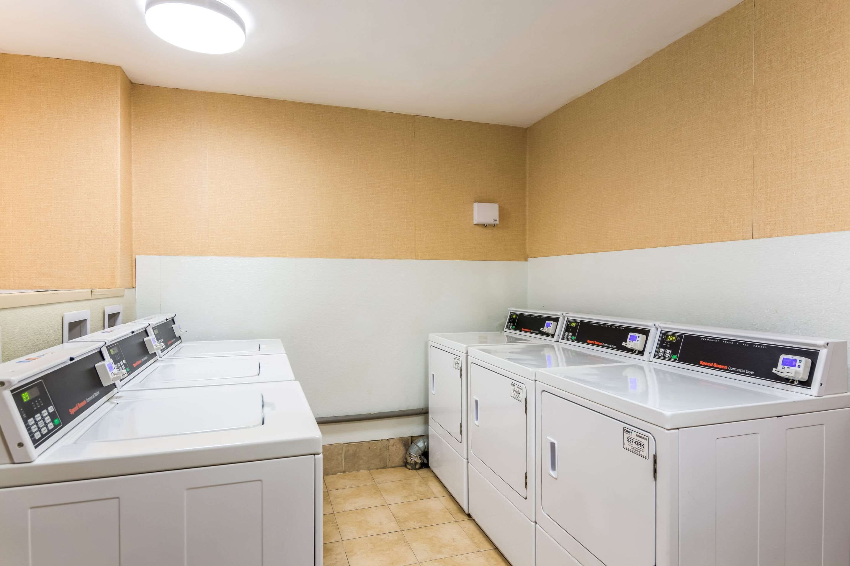 A laundry room with four white front-loading washing machines and dryers, a small ventilation duct, and a light fixture on the ceiling.