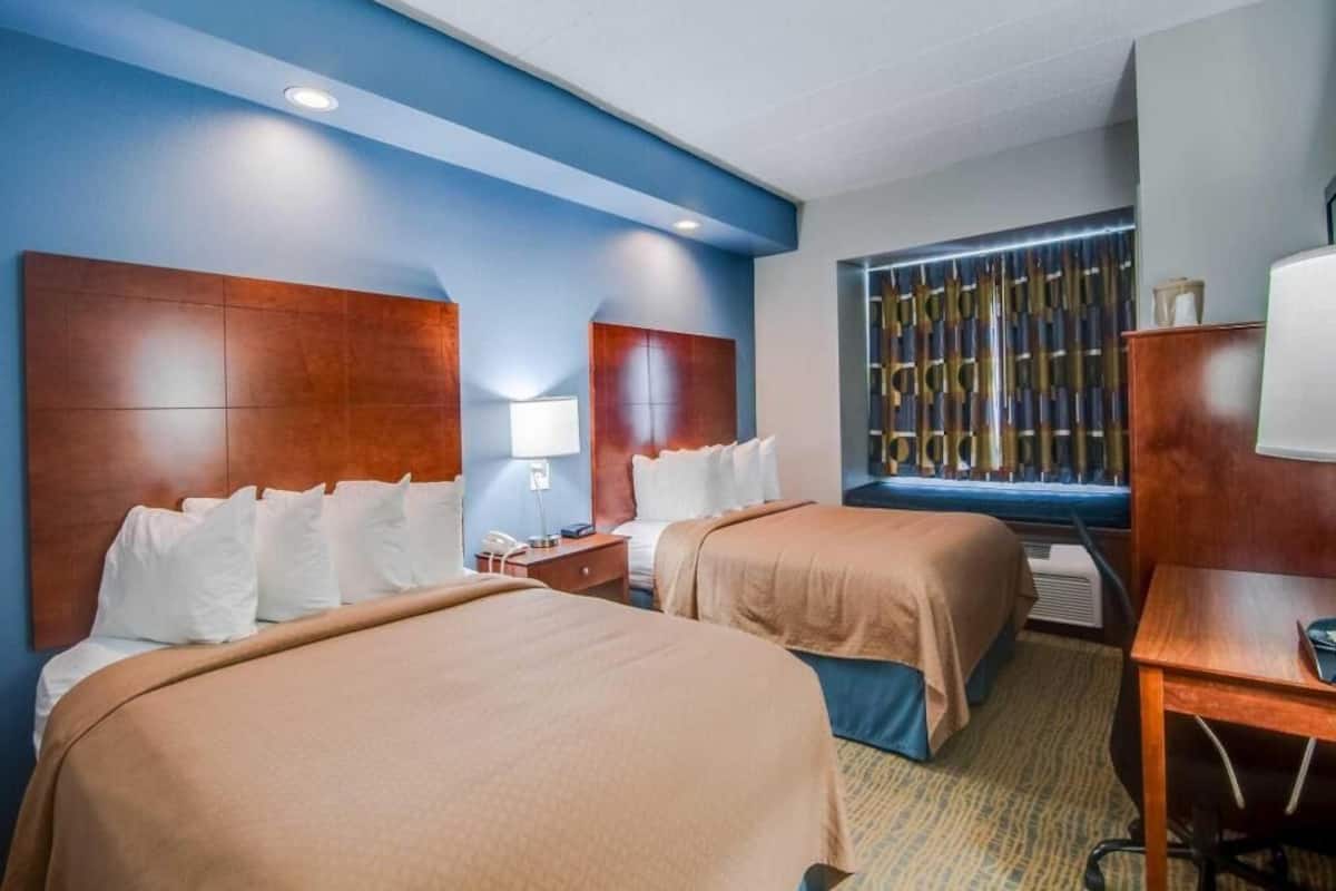 A hotel room with two beds, a desk, a chair, a lamp, and a decorative wall panel.