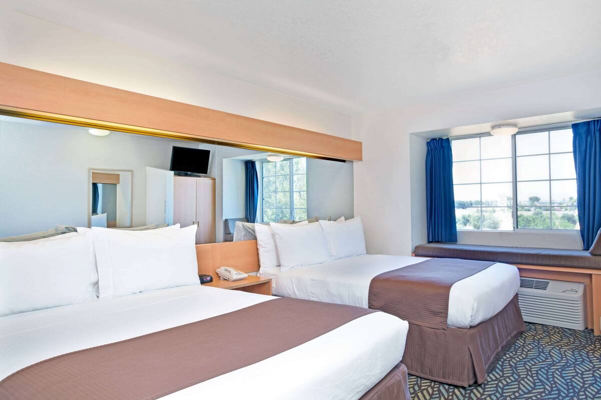 Une chambre d’hôtel avec deux lits, une grande fenêtre avec des rideaux bleus, un téléviseur à écran plat et un bureau avec un téléphone.