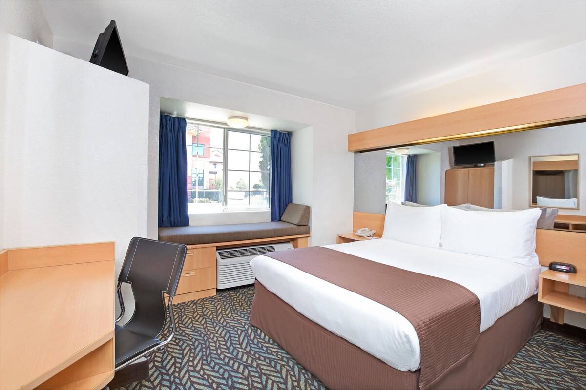 Une chambre d’hôtel comprenant un lit, un bureau, une chaise, une fenêtre avec des rideaux, une télévision et un miroir.