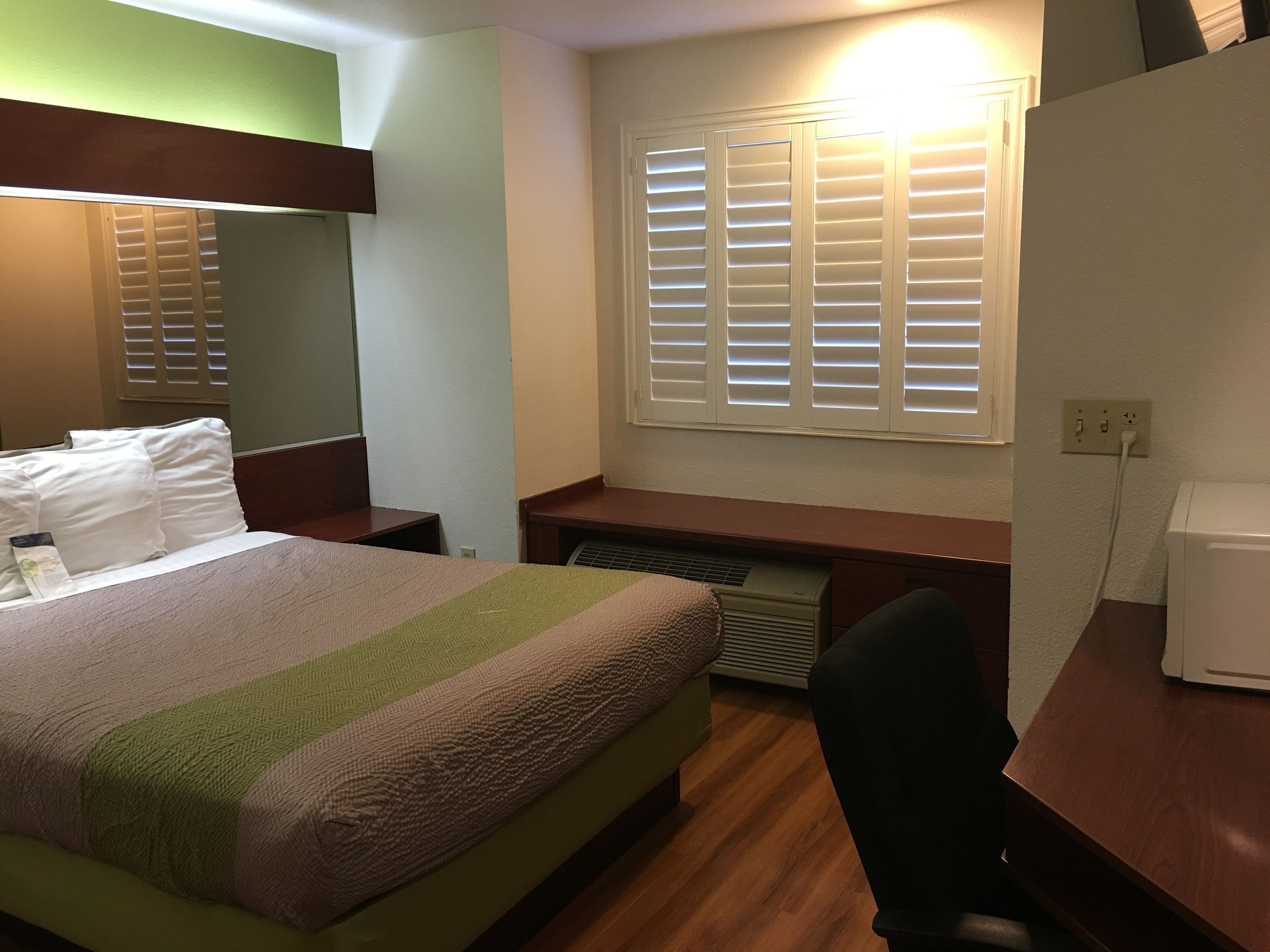 A hotel room with a bed, a desk, a chair, a heater, and a window with blinds.