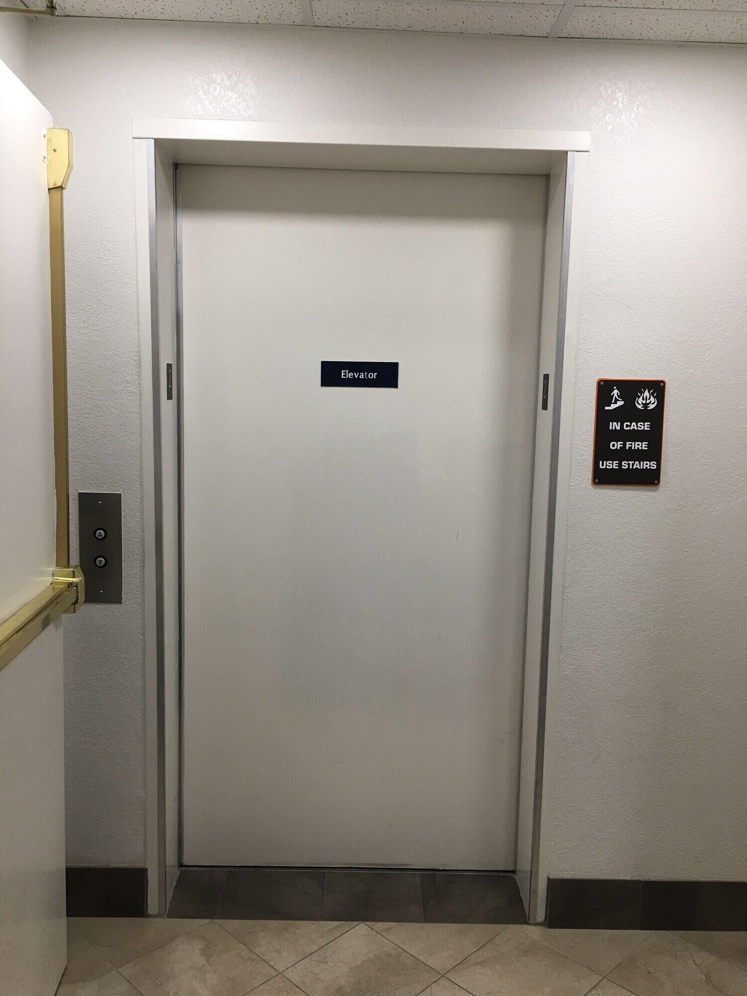 Een liftdeur met een bordje waarop staat 'lift' en een ander bordje dat adviseert de trap te gebruiken in geval van brand.
